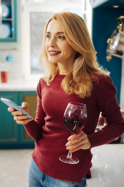 Foto jovem loira e bonita sorrindo e segurando um telefone enquanto bebe um pouco de vinho tinto e pensa