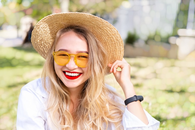 Jovem loira em óculos de sol amarelos, batom vermelho e chapéu de palha está sorrindo amplamente contra a grama verde no parque. Conceito de vibrações positivas de verão, estilo de vida moderno, tempo de sol, close-up