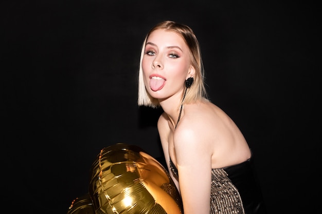 Jovem loira engraçada com balão dourado mostrando a língua enquanto fazia uma careta durante a celebração e curtindo a festa