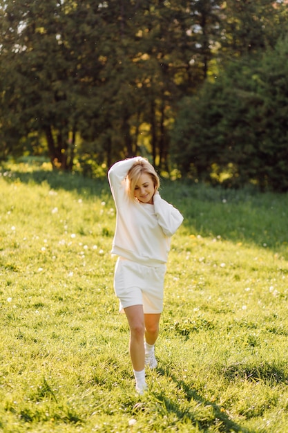 Jovem loira está vestindo um moletom branco, sorrindo e andando na floresta