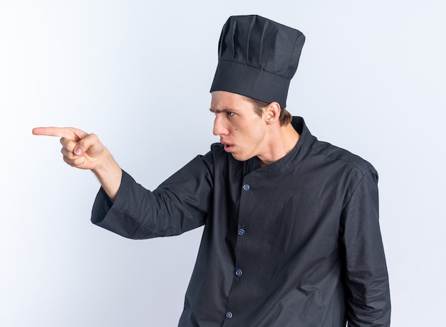 Jovem loira estrita cozinheiro com uniforme de chef e boné olhando e apontando para o lado