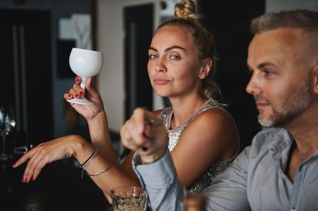 Jovem loira europeia com um copo nas mãos, olhando para a câmera sentada no bar perto de um belo homem branco de meia idade, conversando, se divertindo juntos, flertando, bebendo álcool