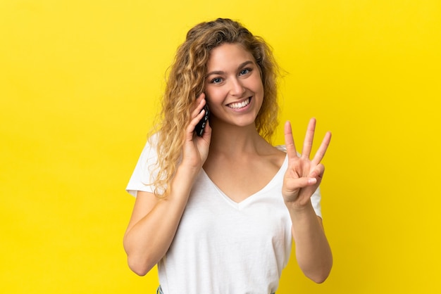 Jovem loira feliz usando telefone celular isolado em um fundo amarelo e contando três com os dedos