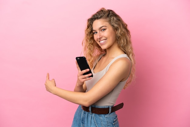 Jovem loira isolada em um fundo rosa usando um telefone celular e apontando para trás