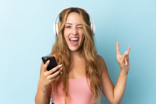 Jovem loira isolada em uma parede azul ouvindo música com um celular fazendo gesto de rock