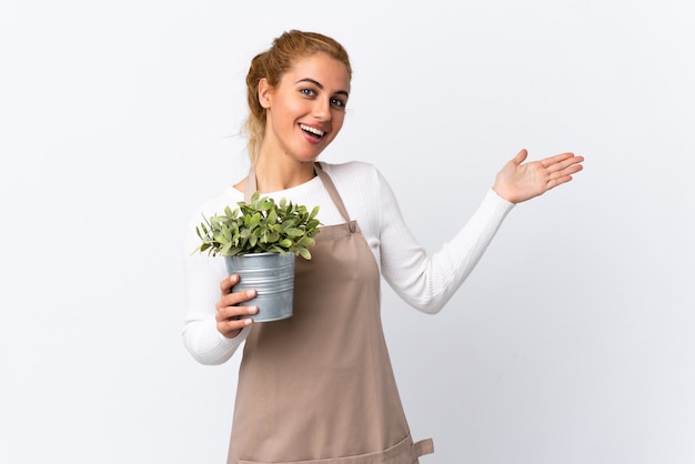 Jovem loira jardineira segurando uma planta sobre uma parede branca isolada, estendendo as mãos para o lado para convidar para vir