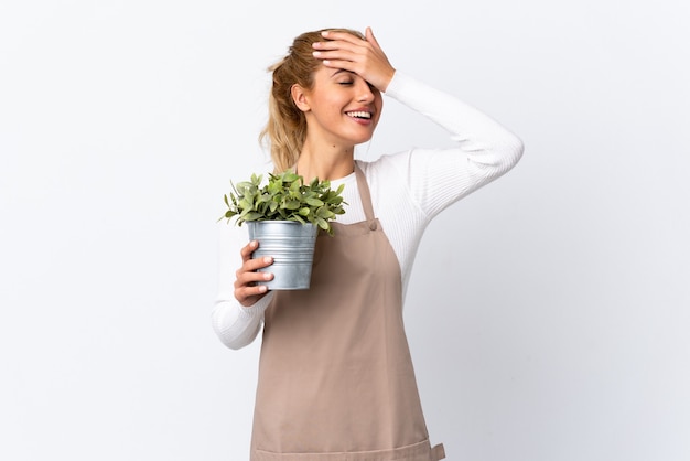 Jovem loira jardineira segurando uma planta sobre uma parede branca isolada rindo