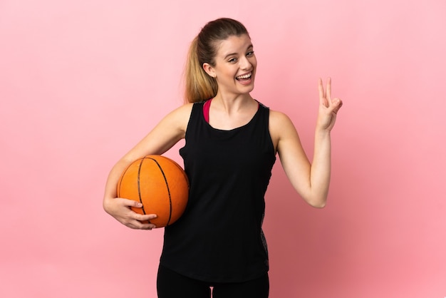 Jovem loira jogando basquete isolada na parede rosa sorrindo e mostrando sinal de vitória