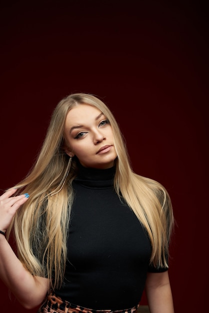 jovem loira linda em preto até os joelhos, sentada em uma cadeira e posando em um fundo vermelho de estúdio