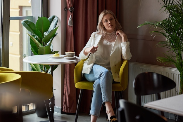 Foto jovem loira linda em uma jaqueta branca senta-se em um café e posa com um rosto positivo, em cima da mesa é uma xícara de café e bolo