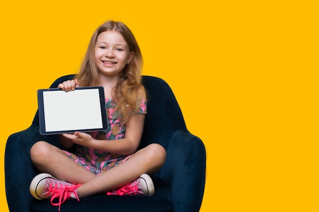 Jovem loira mostrando para a câmera um tablet digital com tela branca sorrindo em uma parede amarela do estúdio com espaço livre