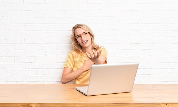 jovem loira no laptop sorrindo com uma mão ao lado do coração e a outra esticada na frente