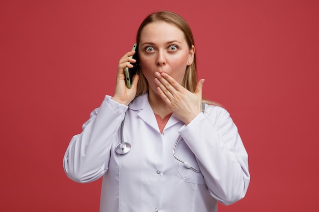 Foto jovem loira preocupada com uma médica usando um manto médico e um estetoscópio pendurado no pescoço falando no telefone e fazendo um gesto de opa