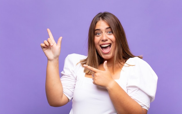 Jovem loira se sentindo alegre e surpresa, sorrindo com uma expressão chocada e apontando para o lado