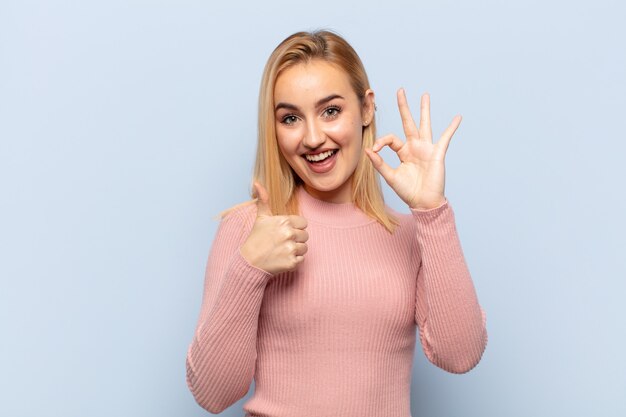 Jovem loira se sentindo feliz, maravilhada, satisfeita e surpresa, mostrando gestos de ok e polegar para cima, sorrindo