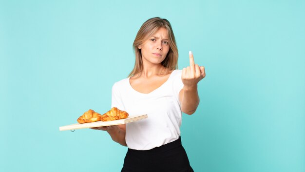 Jovem loira se sentindo irritada, irritada, rebelde e agressiva segurando uma bandeja de croissant