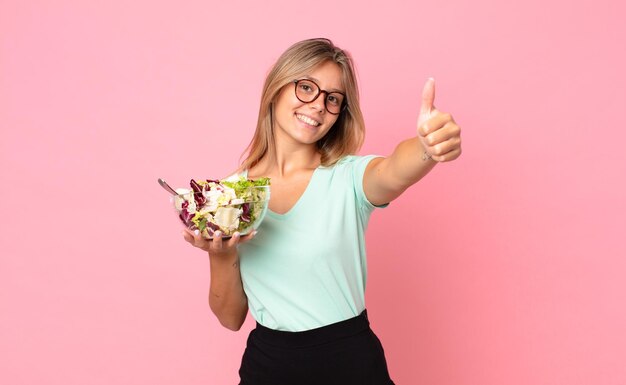 Jovem loira se sentindo orgulhosa, sorrindo positivamente com o polegar para cima e segurando uma salada