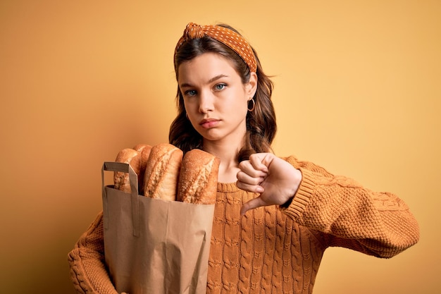 Jovem loira segurando o saco de papel do supermercado cheio de pão fresco sobre fundo amarelo com sinal negativo de rosto zangado, mostrando desagrado com o conceito de rejeição de polegares para baixo