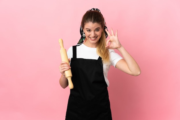 Jovem loira segurando um rolo de massa isolado em um fundo rosa e mostrando um sinal de ok com os dedos