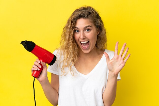 Jovem loira segurando um secador de cabelo isolado em um fundo amarelo e contando cinco com os dedos