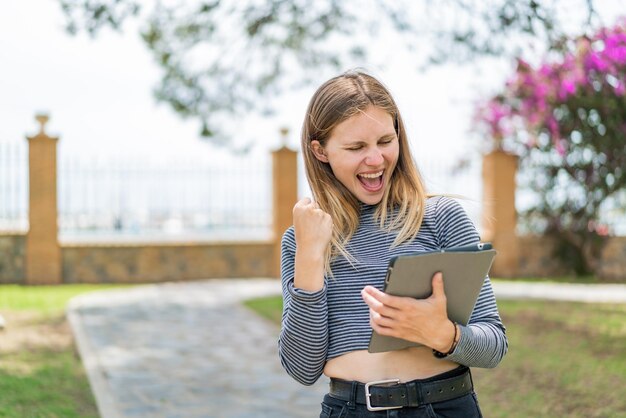 Jovem loira segurando um tablet ao ar livre comemorando uma vitória