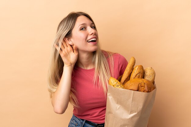 Jovem loira segurando uma sacola cheia de pães isolada em bege ouvindo algo