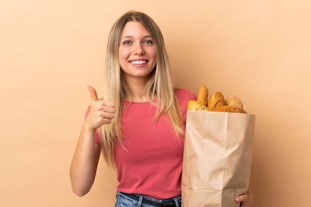 Jovem loira segurando uma sacola cheia de pães isolada na parede bege e fazendo um gesto de polegar para cima