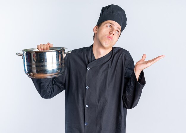 Jovem loira sem noção do cozinheiro com uniforme de chef e boné segurando a panela, olhando para cima, mostrando a mão vazia, isolada na parede branca