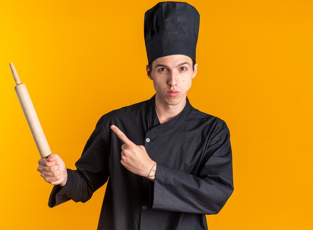 Jovem loira séria cozinheiro em uniforme de chef e boné segurando e apontando para o rolo de massa