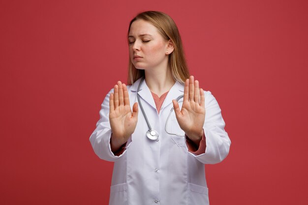 Jovem loira séria usando roupão médico e estetoscópio no pescoço, fazendo gesto de parada com os olhos fechados