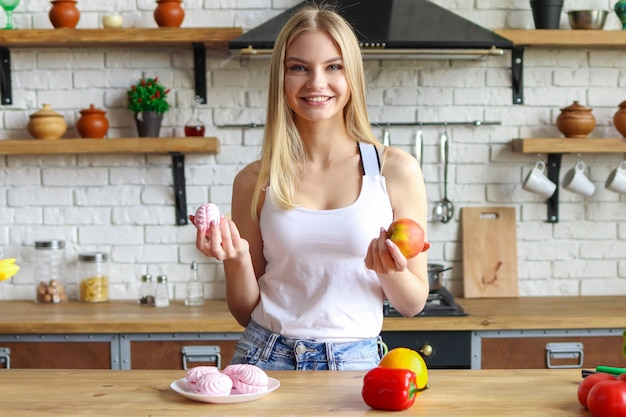 Jovem loira sorri, mulher na cozinha escolhe entre doces e frutas, alimentos saudáveis