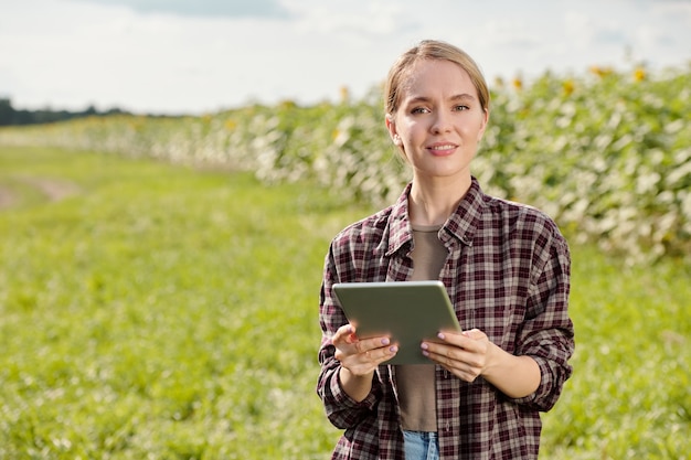 Jovem loira sorridente agricultora em trajes de trabalho, olhando para você em pé na frente da câmera e usando tablet digital contra um campo verde