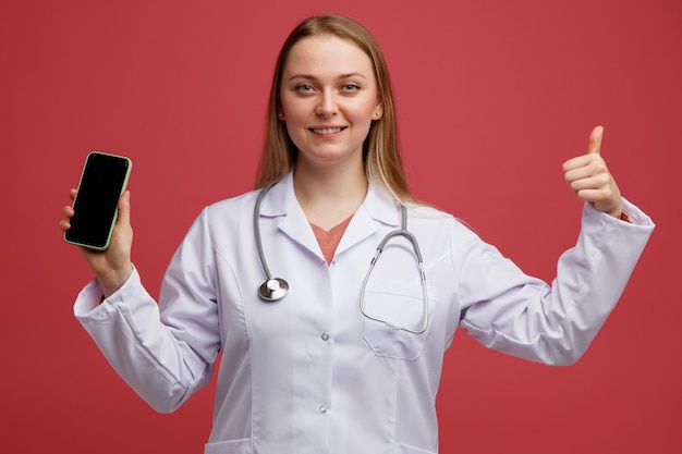 Jovem loira sorridente com um robe médico e um estetoscópio pendurado no pescoço, segurando um telefone celular e mostrando o polegar