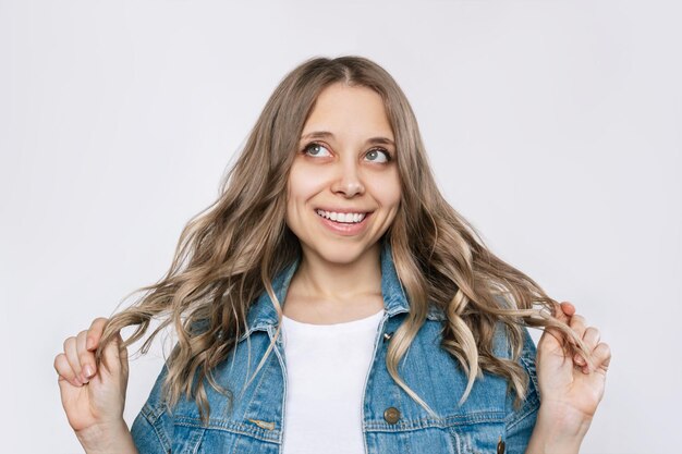 Jovem loira sorridente imagina ou pensa segurando seus fios de cabelo ondulado e olhando para cima