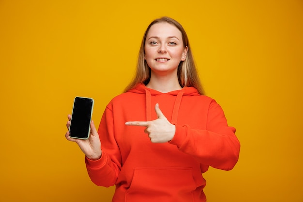 Jovem loira sorridente segurando e apontando para o celular