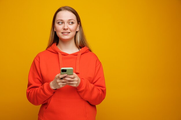 Jovem loira sorridente segurando um telefone celular com as duas mãos, olhando para o lado