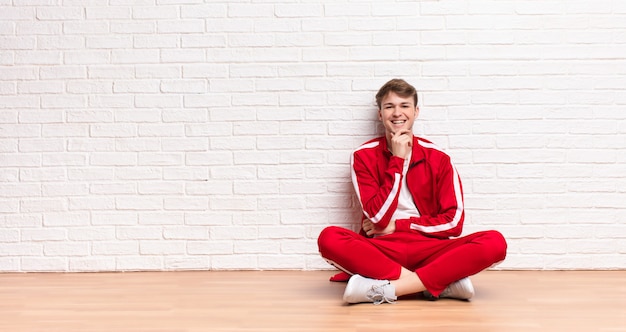 Jovem loira sorrindo, curtindo a vida, sentindo-se feliz, amigável, satisfeito e despreocupado com a mão no queixo, sentado no chão