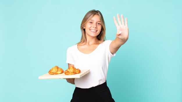 jovem loira sorrindo e parecendo amigável, mostrando o número quatro e segurando uma bandeja de croissant