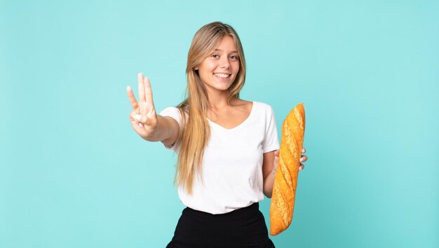 Jovem loira sorrindo e parecendo amigável, mostrando o número três e segurando uma baguete de pão