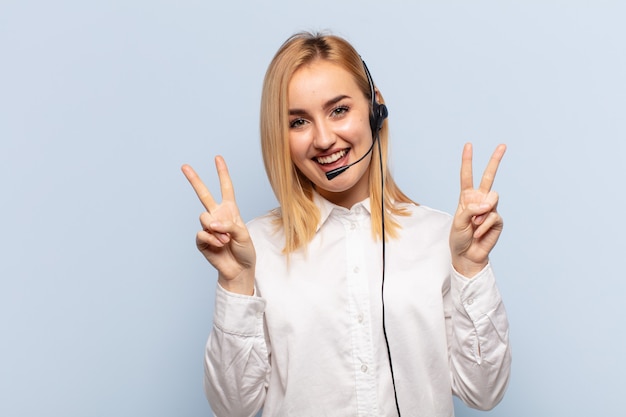 Jovem loira sorrindo e parecendo feliz, amigável e satisfeita, gesticulando vitória ou paz com as duas mãos