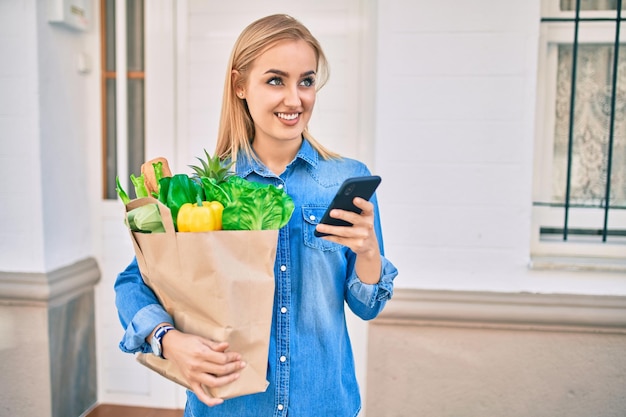Jovem loira sorrindo feliz usando smartphone e segurando saco de papel de mantimentos na cidade