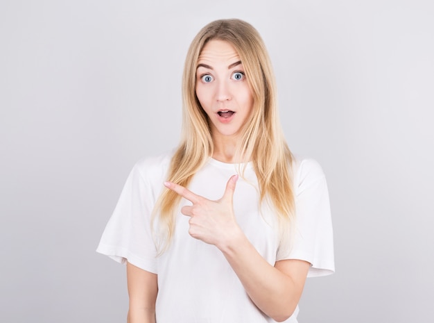 Jovem loira surpresa aponta o dedo para o espaço à sua esquerda. Retrato de uma menina surpresa em um fundo cinza.