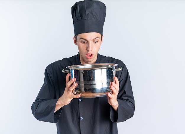 Jovem loira surpresa surpresa com uniforme de chef e boné segurando e olhando para dentro do pote isolado na parede branca