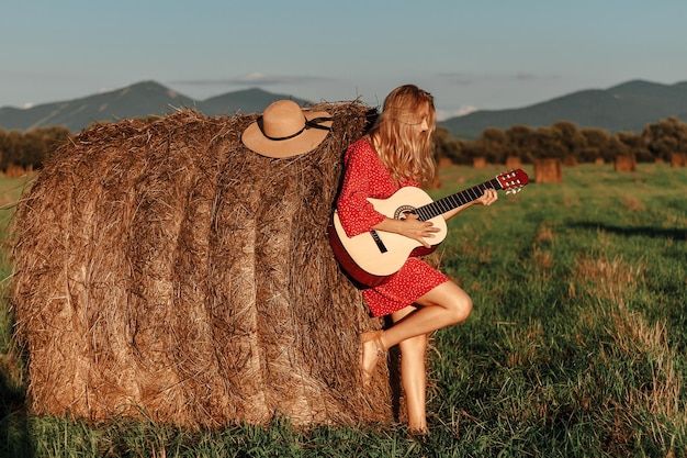 Jovem loira toca violão perto de um palheiro em um campo por do sol