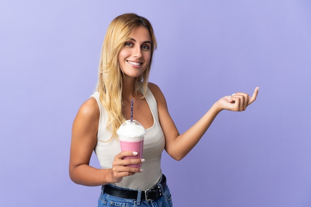 Jovem loira uruguaia com milk-shake de morango isolado na parede roxa estendendo as mãos para o lado para convidar para vir