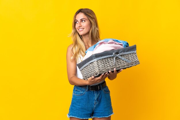 Jovem loira uruguaia segurando uma cesta de roupas isolada na parede amarela olhando para o lado e sorrindo