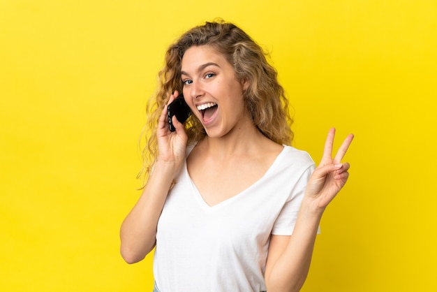 Jovem loira usando telefone celular isolado em um fundo amarelo, sorrindo e mostrando sinal de vitória