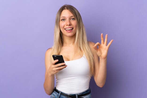 Jovem loira usando telefone celular isolado na parede roxa e mostrando um sinal de ok com os dedos