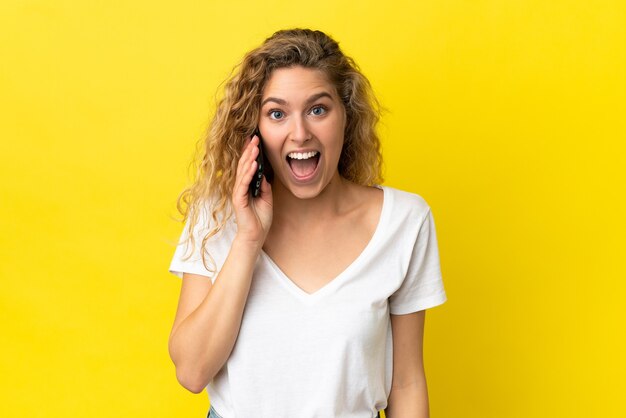 Jovem loira usando um celular isolado em um fundo amarelo com uma expressão facial surpresa