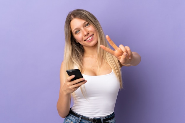 Jovem loira usando um celular isolado na parede roxa sorrindo e mostrando o sinal da vitória